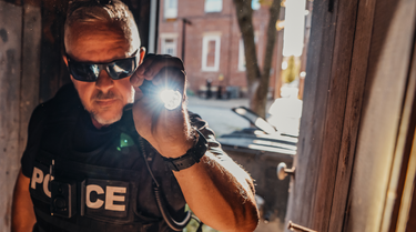 Image of a white male police officer holding a tactical flashlight while entering a dark building during the day. 